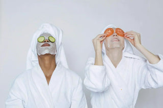 Two women in bathrobes enjoying a relaxing spa day with sheet masks and fresh cucumber and tomato slices for skincare.