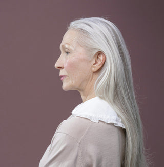 Side profile of an older woman with long grey hair, wearing a soft pink top, symbolising natural ageing and graceful skin confidence.