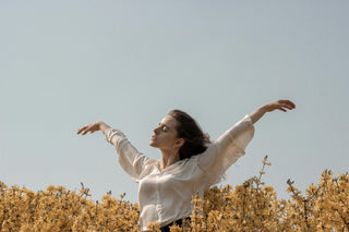 Woman standing in a field of yellow flowers with arms raised and eyes closed, expressing calm and freedom under a clear sky