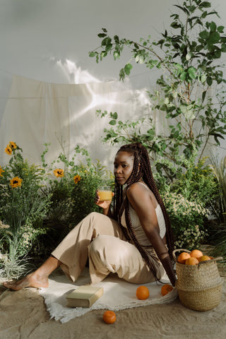 Woman holding glass of juice sat on the floor surrounded by greenery and peaches.