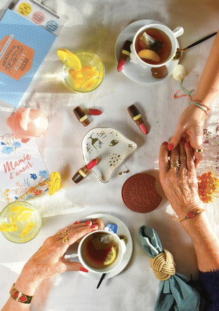 A warm tea-time scene with two hands holding across generations, surrounded by tea, chocolate treats, and a "Mamie d'amour" card, symbolising love and connection.