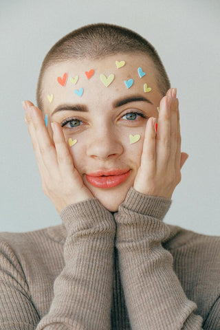 A young person with a shaved head and bright eyes smiles gently, cupping their face with both hands.