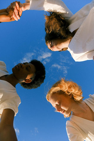 A group of three people in white outfits hold hands while standing in a circle, viewed from below against a bright blue sky