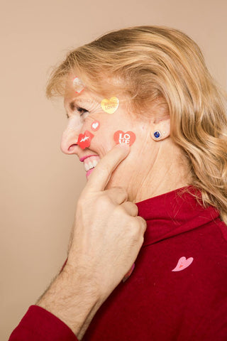 a smiling woman with blonde hair wearing a red turtleneck. Her face is decorated with colorful heart-shaped stickers with words like "LOVE" and "REAL LOVE