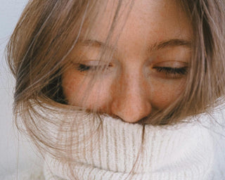 A cosy close-up of a woman wrapped in a thick white knit sweater, her face partially covered by the collar