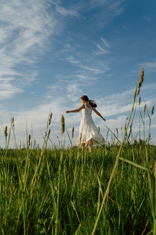 A woman in a white dress joyfully dances in a sunlit meadow against a clear blue sky.