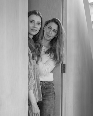 A black and white photograph of two women standing behind a wooden door frame, wearing casual knit and denim clothing.